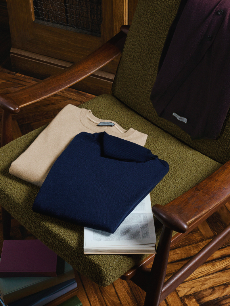 Maglieria da uomo Canali: una maglia beige girocollo e una polo in maglia blu piegate su una poltrona verde, accanto a un libro aperto, in un ambiente dai toni caldi e raffinati.
