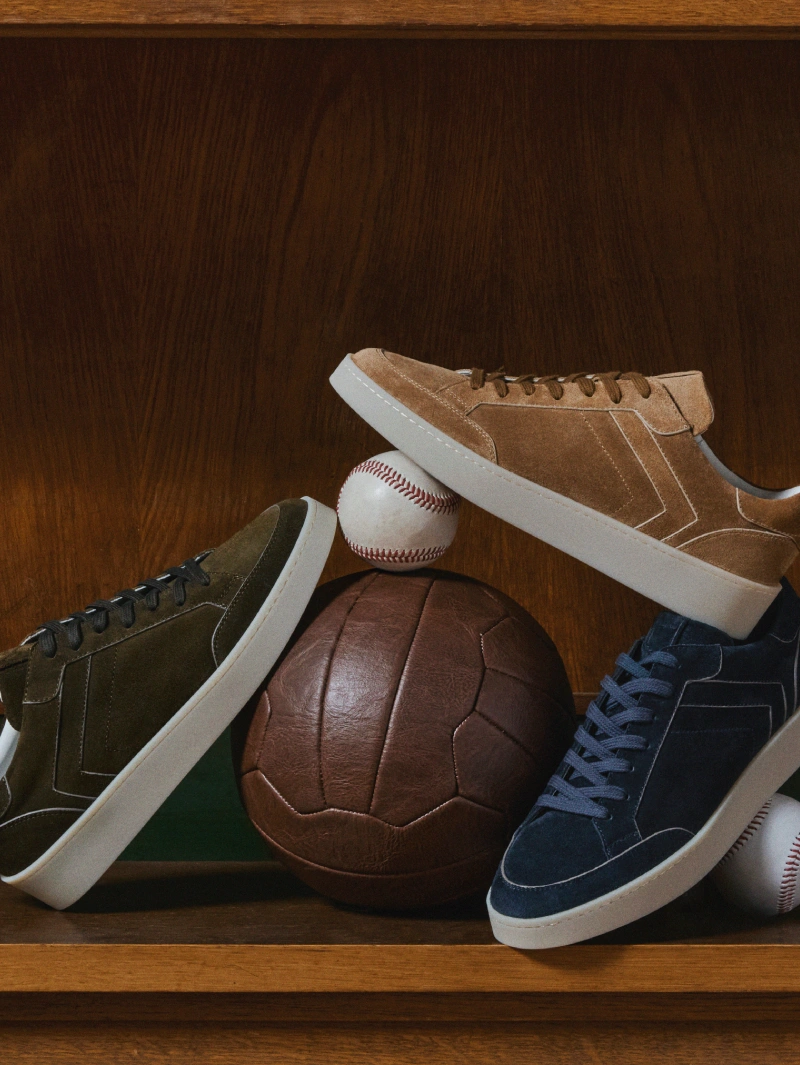Fall/Winter 2026 suede sneakers in olive green, tan and navy blue with white rubber soles, displayed on a wooden shelf with vintage football and baseball accessories.