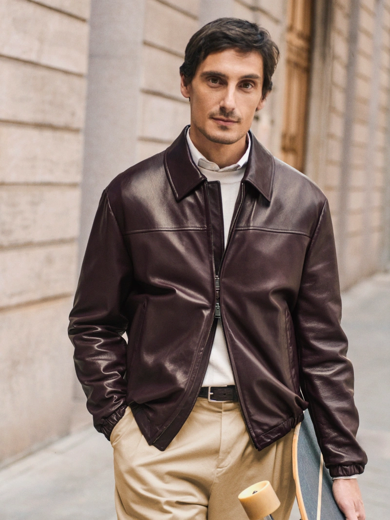 Fall/Winter 2026 look featuring a burgundy leather jacket, light knitwear and beige trousers, styled with a skateboard against a classic city backdrop.
